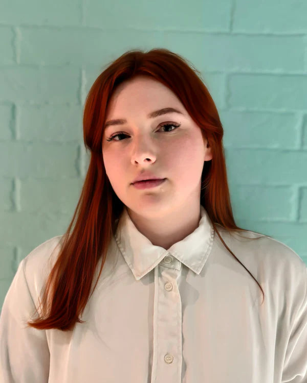 Person with red hair wearing a light-colored shirt against a teal brick wall.